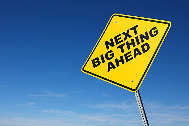 A yellow road sign indicates that the next big thing is ahead in front of a blue sky with wispy clouds at the horizon. The perspective is from a low camera angle. Road sign is placed on right side of image allowing for copy space on the left side of image.
